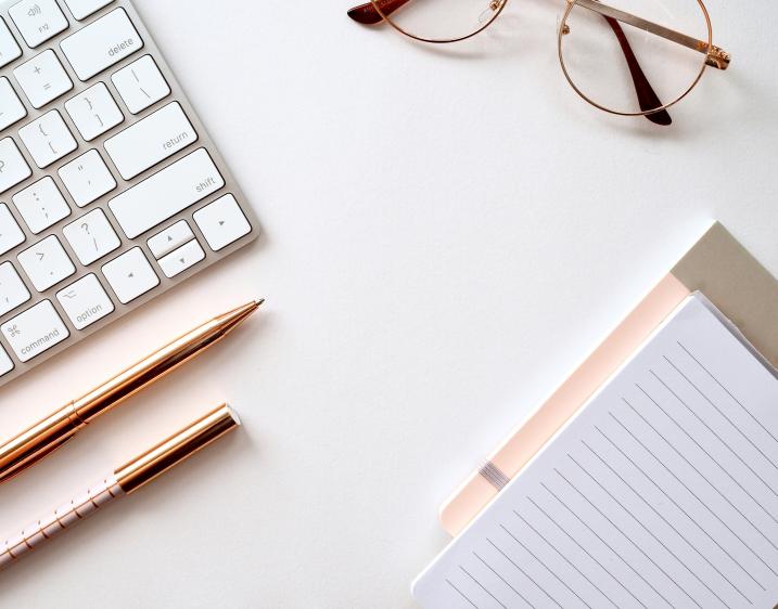 A picture of a keyboard, glasses, pens, and notepad laying on a desk